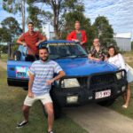 Five people pose in front of a blue 4WD vehicle parked on a paved path with trees and a building in the background.