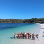 A group of people stand in shallow water on a white sand beach, raising their arms, with turquoise water and a forested shoreline under a clear blue sky.