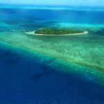 A small, lush green island surrounded by turquoise shallow waters and coral reefs, set in a vast blue ocean under a clear sky.