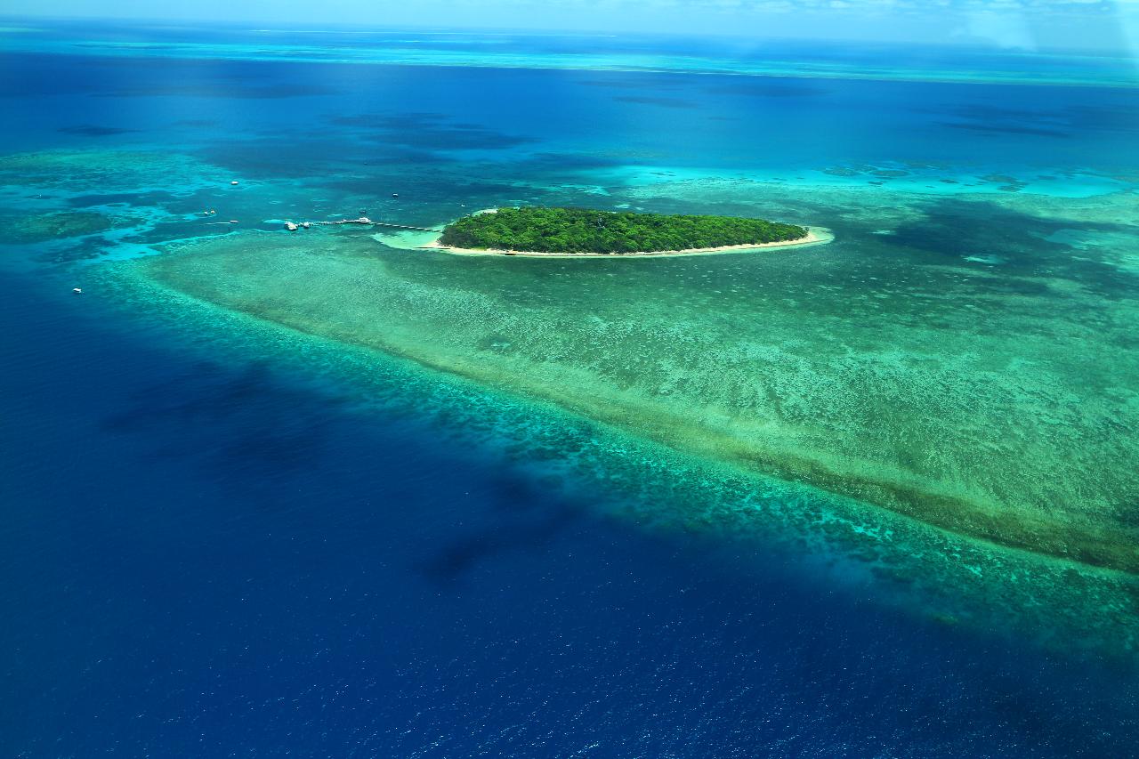 A small, lush green island surrounded by turquoise shallow waters and coral reefs, set in a vast blue ocean under a clear sky.