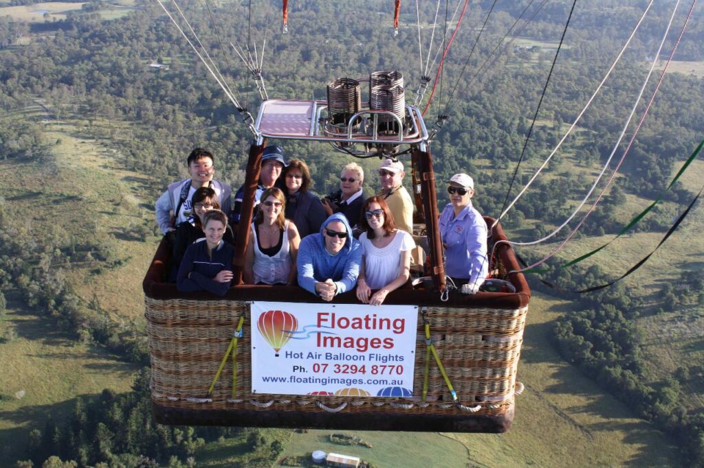 A group of people in a hot air balloon basket with a "Floating Images Hot Air Balloon Flights" sign, flying over a green landscape.