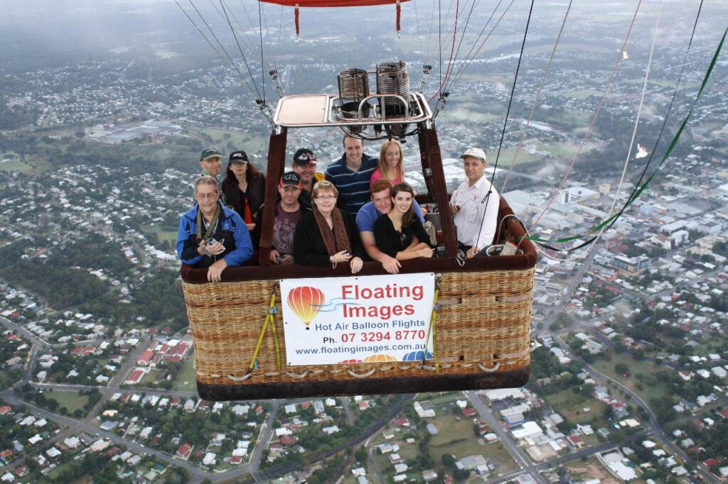 A group of people stand in a hot air balloon basket above a town, with a "Floating Images" sign displaying contact details on the basket.