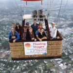 A group of people stand in a hot air balloon basket above a town, with a "Floating Images" sign displaying contact details on the basket.