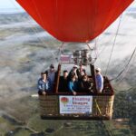 A group of people ride in a hot air balloon basket high above a rural landscape with fog and farmlands visible below.