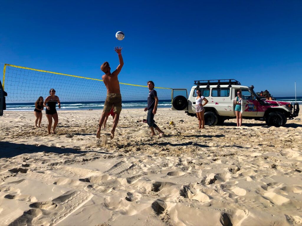 A group of people play beach volleyball on sand near the ocean, with a volleyball net and a parked white SUV in the background under a clear blue sky.