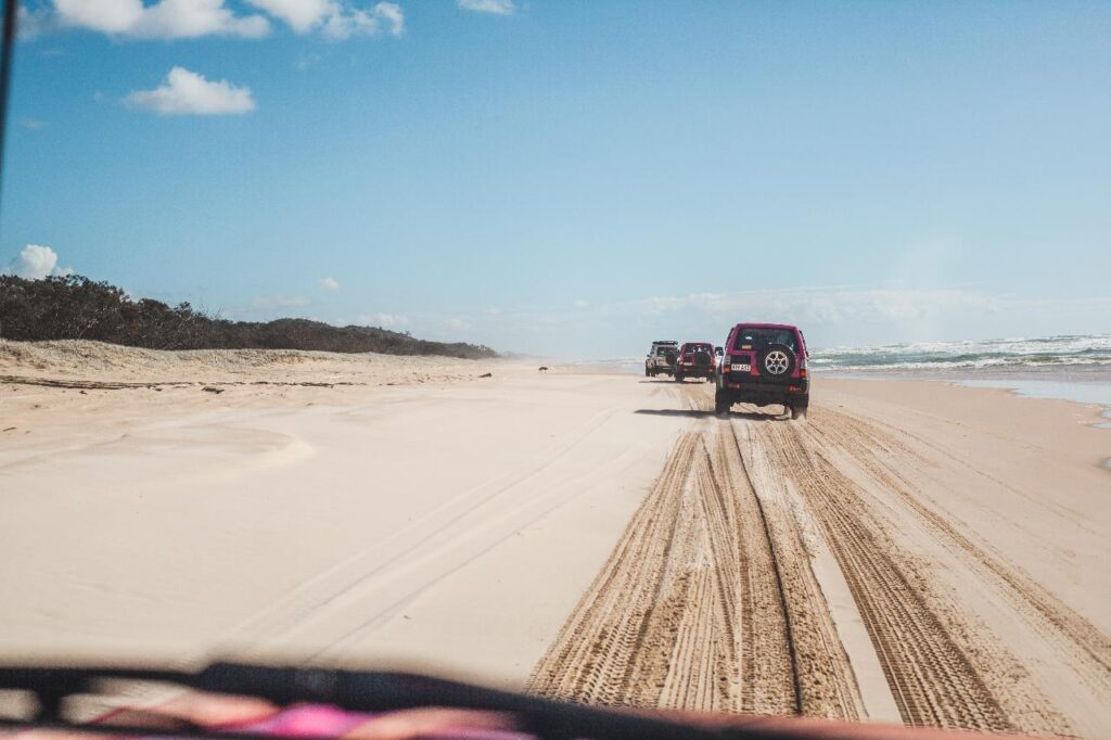 Three SUVs drive along sandy tire tracks on a wide beach under a blue sky, with the ocean on the right and vegetation on the left.