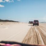 Three SUVs drive along sandy tire tracks on a wide beach under a blue sky, with the ocean on the right and vegetation on the left.