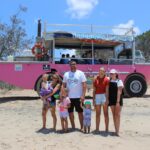 A group of people stand on a sandy beach in front of a pink amphibious tour vehicle with trees and blue sky in the background.