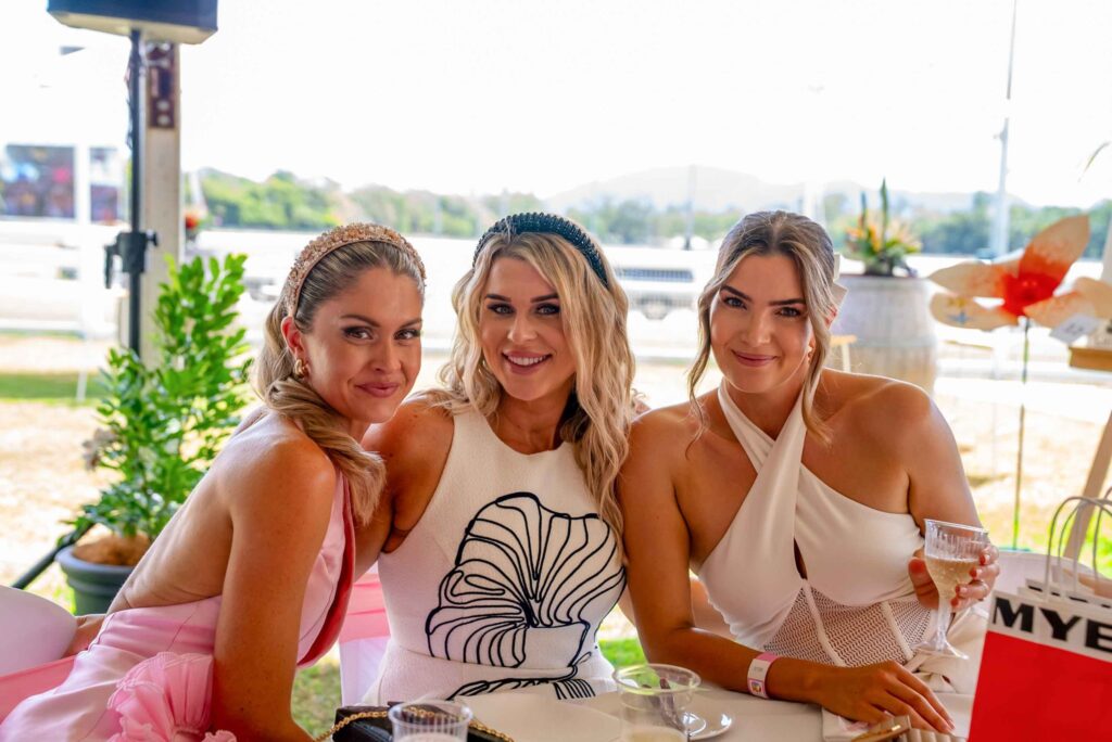 Three women sit at a table outdoors, dressed in light-colored, stylish outfits, smiling at the camera while holding drinks.