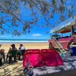 A group of people sit on plastic chairs under tree shade by the beach near a pink off-road vehicle, with trays and drinks on a table in the foreground.