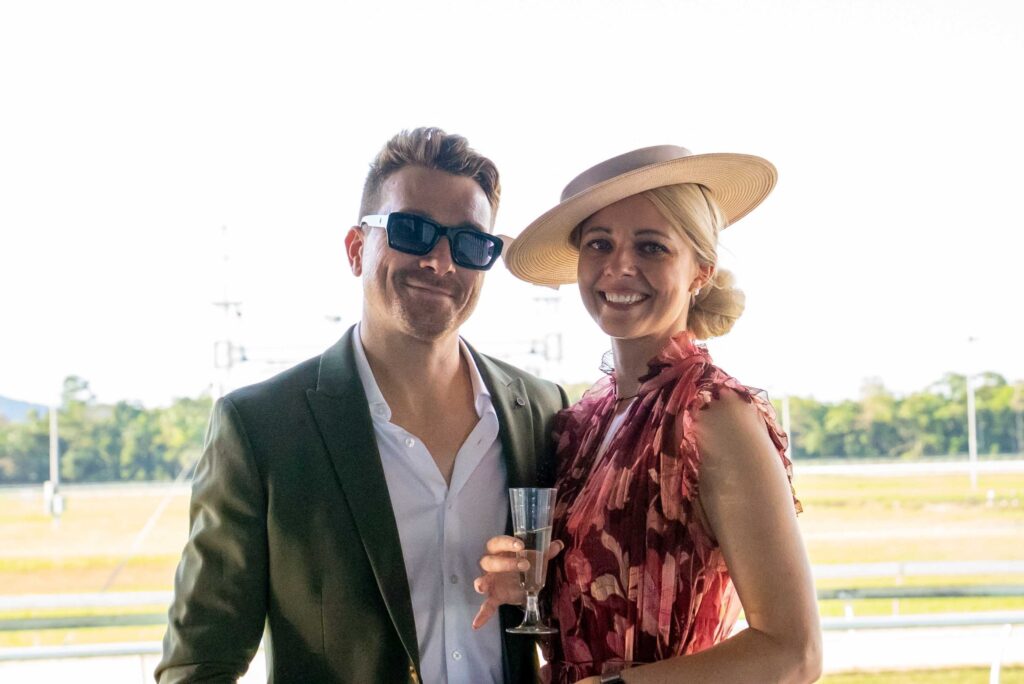 A man in sunglasses and a green suit stands next to a smiling woman in a pink dress and wide-brimmed hat holding a champagne glass at an outdoor event.
