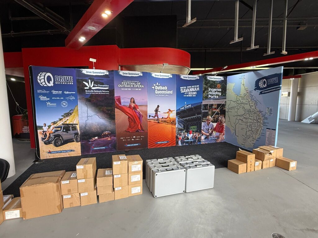 A display booth with large tourism banners, cardboard boxes, and silver cases set up in an indoor venue with a concrete floor.