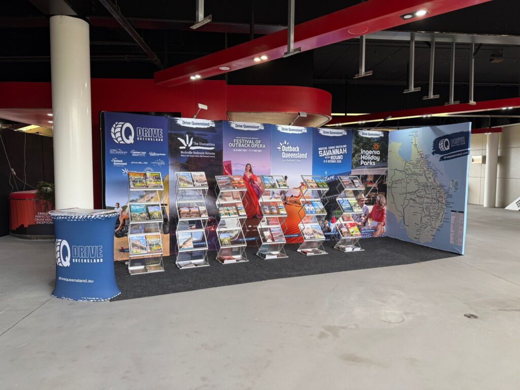 A tourism information booth with brochures on display racks, promotional banners, and a large map, set up indoors on a black carpet.