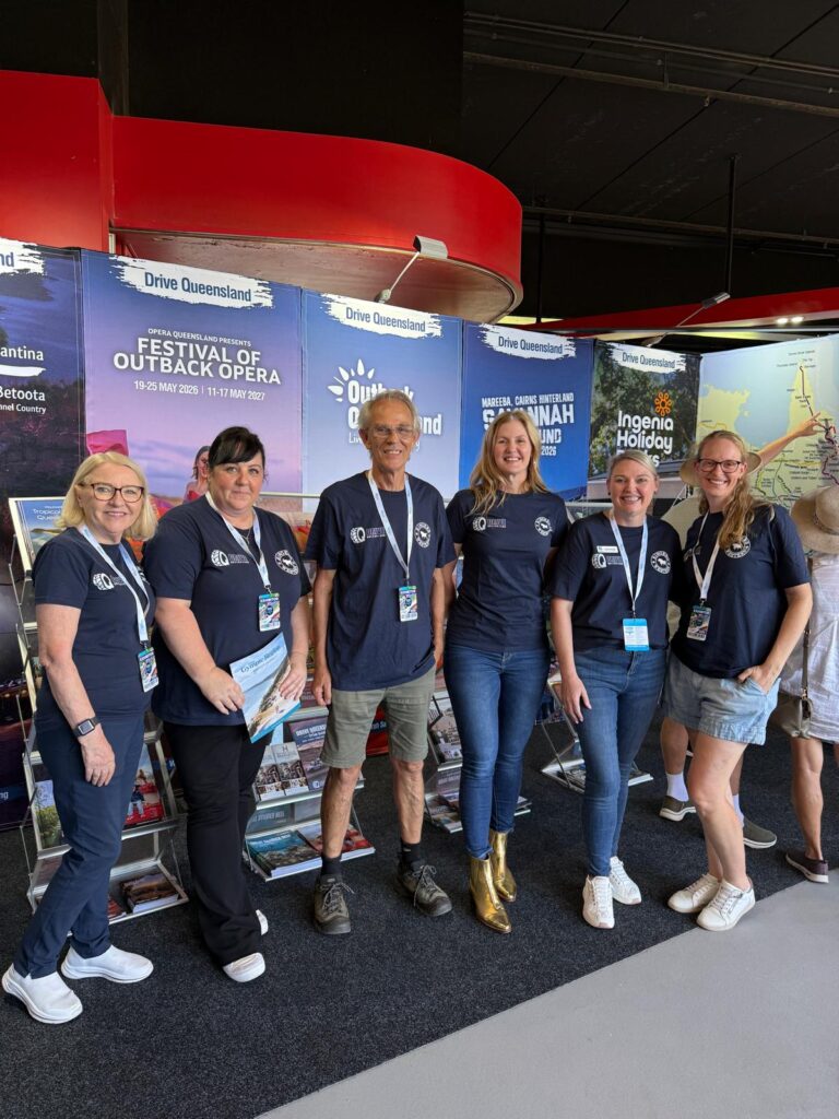 Six people wearing matching navy shirts pose in front of a display promoting Queensland tourism events and destinations at an indoor venue.