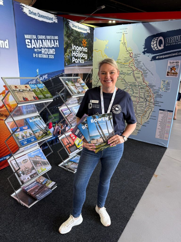 A woman stands in front of a tourism booth holding brochures, with maps, pamphlets, and travel posters for Queensland, Australia on display behind her.