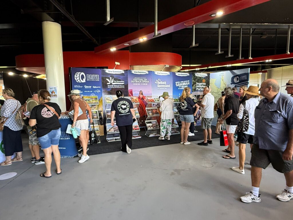 People stand in line at an indoor tourist information booth with brochures and banners displaying local attractions and destinations.