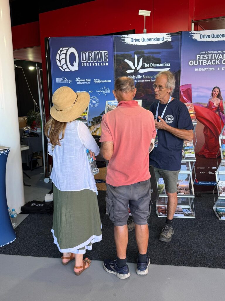 Three people are standing and talking in front of a tourism information booth with banners and brochures for Drive Queensland and regional attractions.