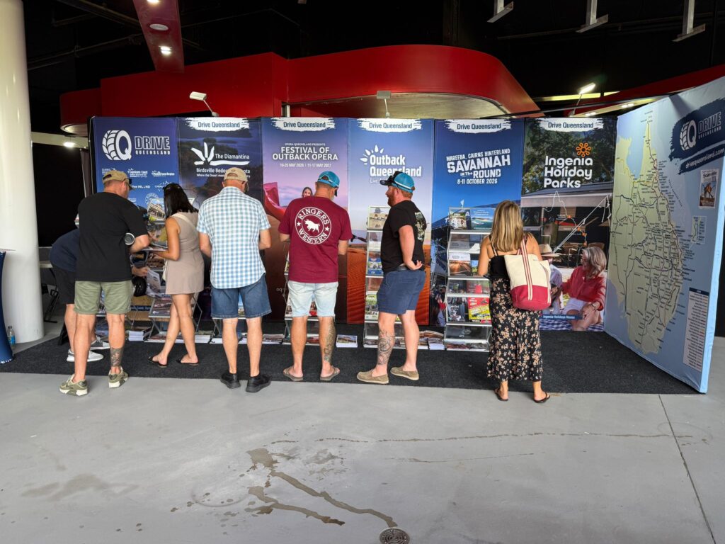 A group of people stand at a tourism booth with promotional banners and brochures about Queensland attractions and holiday parks.