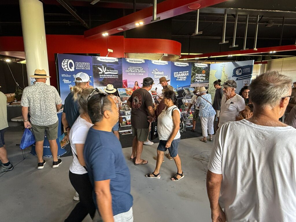 People walk and browse at an indoor exhibition booth with brochures and posters displayed on the back wall.