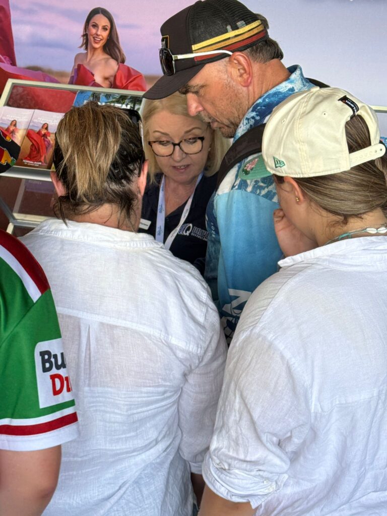Four people stand closely together, facing a woman who appears to be explaining something. The group is indoors and colorful photos are displayed on the wall behind them.