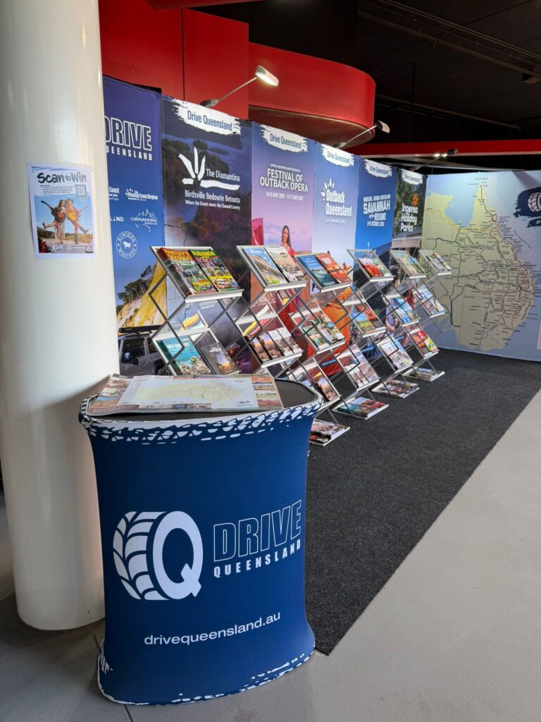 A Drive Queensland display booth features travel brochures, a map of Queensland, and promotional posters at an indoor event.