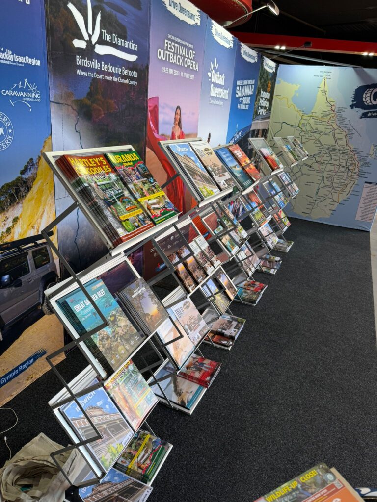 A display of travel brochures and magazines arranged on racks at a tourism information booth, with posters and a large map on the wall behind.
