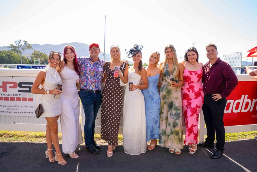 A group of eight people, dressed in colorful dresses and shirts, pose together outdoors, smiling and holding drinks at a racetrack event.