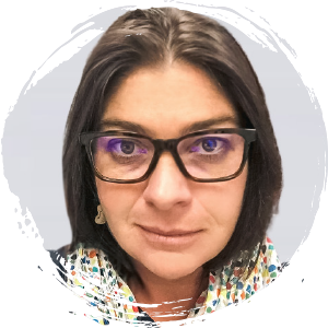 A woman with shoulder-length brown hair and glasses looks at the camera. She is wearing a patterned scarf and a light-colored top, with a neutral background behind her.