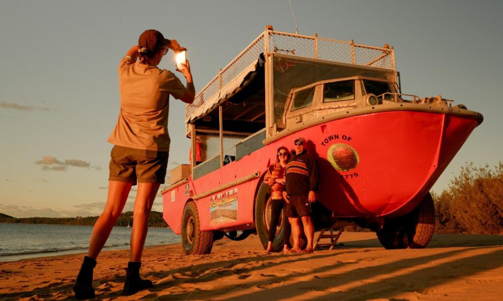 Person taking a photo of a group standing beside an amphibious tour vehicle on a sandy beach at sunset.