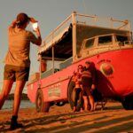 Person taking a photo of a group standing beside an amphibious tour vehicle on a sandy beach at sunset.