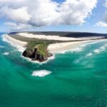 Aerial view of a sandy coastline with turquoise ocean waves, rocky headland, white sand dunes, and partly cloudy sky.