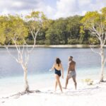 A woman and a man in swimsuits walk barefoot on white sand by a lake, with trees and forest in the background under a partly cloudy sky.