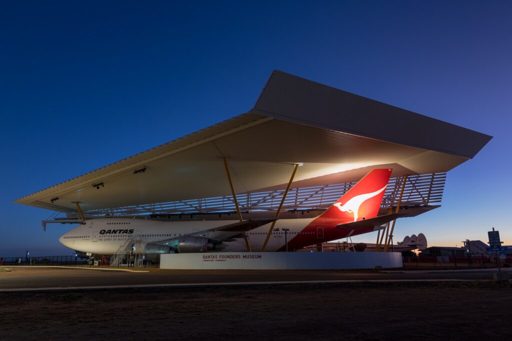 A Qantas Boeing 747 is displayed under a large metal canopy at the Qantas Founders Museum during twilight.