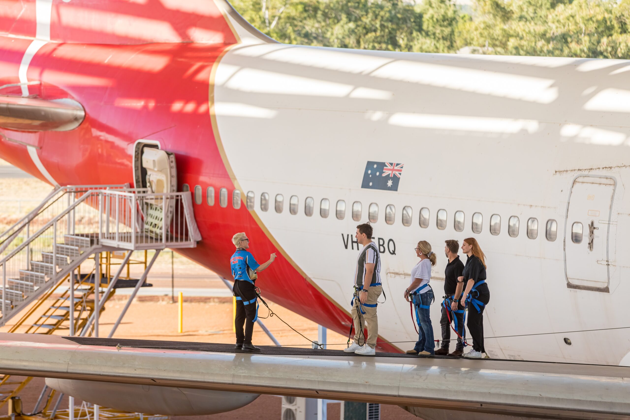 A group of people wearing harnesses stand on the wing of a large airplane near an open passenger door, with a guide speaking to them.