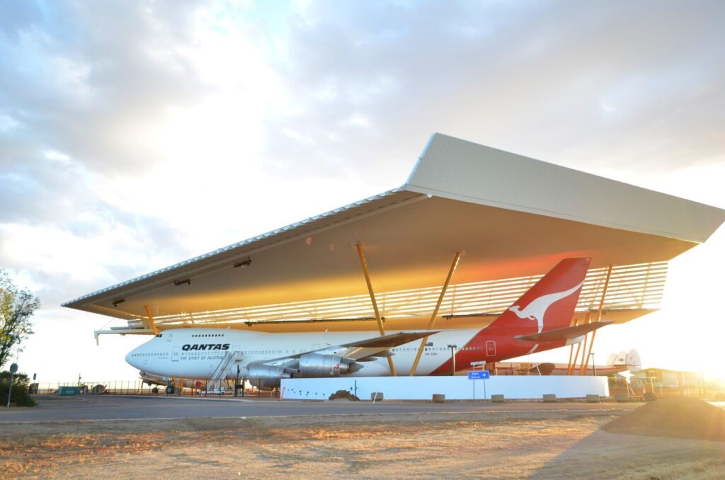 A Qantas Boeing 747 is displayed under a large, modern canopy structure at the Qantas Founders Museum during sunset.