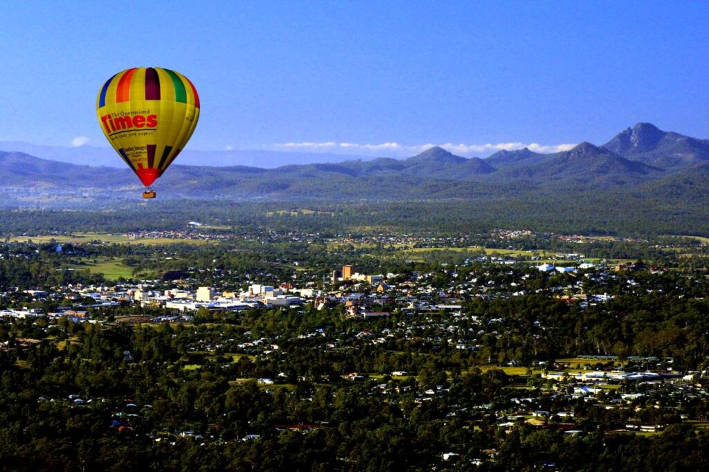 A colorful hot air balloon floats above a town surrounded by greenery and mountains under a clear blue sky.
