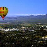 A colorful hot air balloon floats above a town surrounded by greenery and mountains under a clear blue sky.