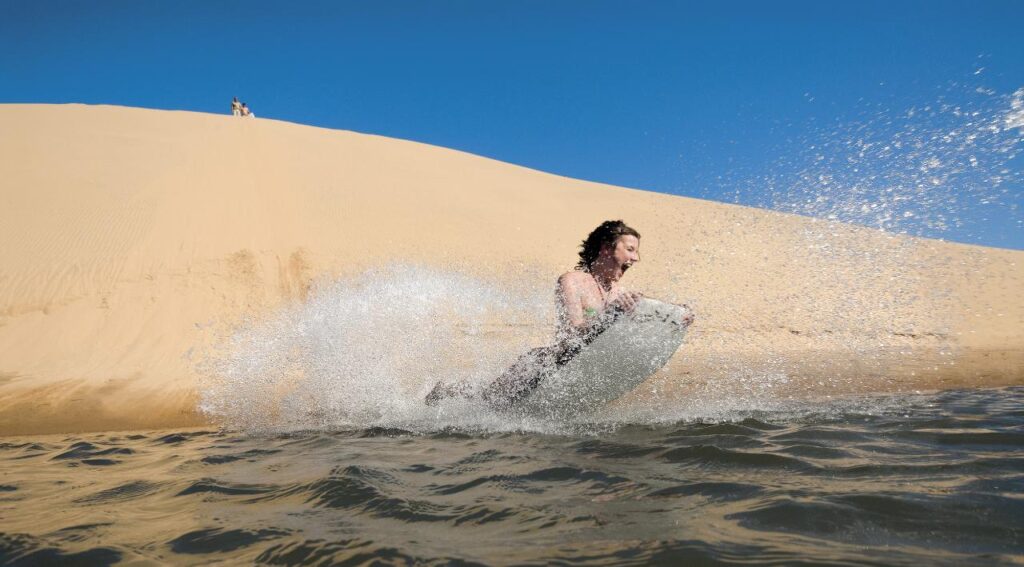 A person rides a bodyboard down a sandy dune and splashes into the water under a clear blue sky.