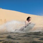 A person rides a bodyboard down a sandy dune and splashes into the water under a clear blue sky.