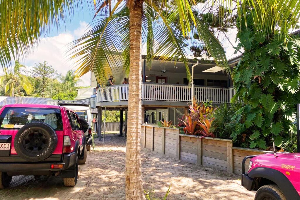 A raised house with a wraparound porch is surrounded by tropical plants. Two magenta vehicles are parked on a sandy driveway in front of the house.