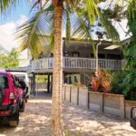 A raised house with a wraparound porch is surrounded by tropical plants. Two magenta vehicles are parked on a sandy driveway in front of the house.