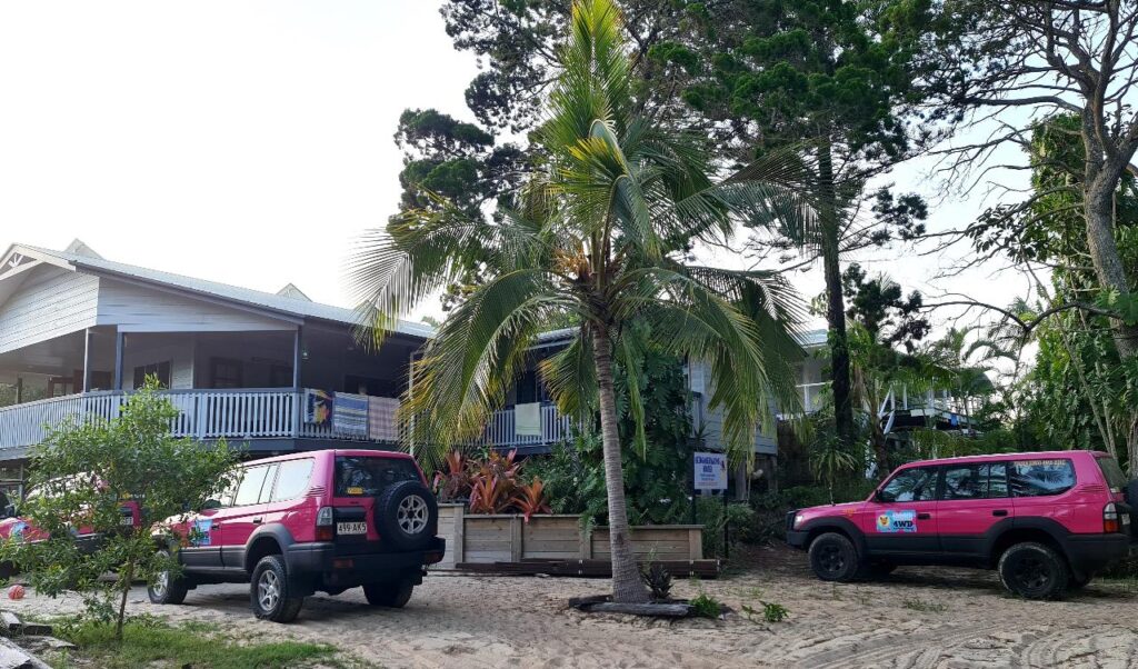 Two pink off-road vehicles are parked on a sandy area in front of a raised white building with a large veranda, surrounded by palm trees and greenery.