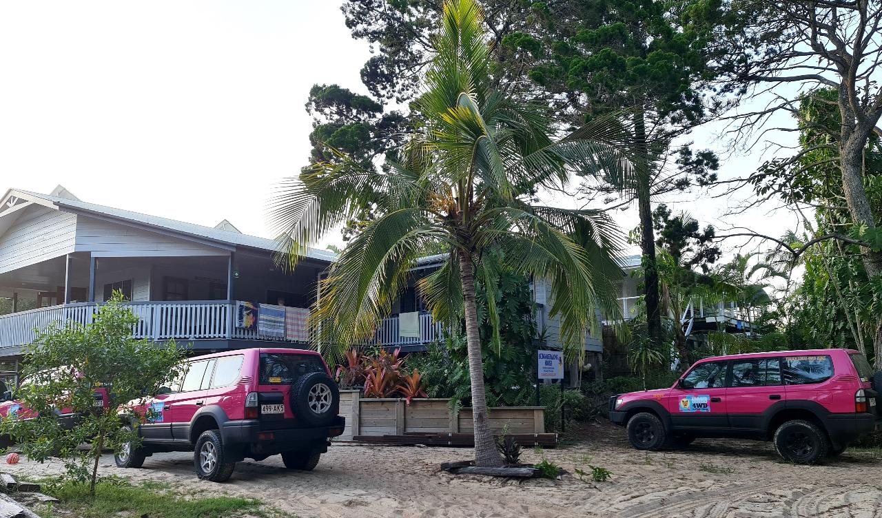 Two pink off-road vehicles are parked on a sandy area in front of a raised white building with a large veranda, surrounded by palm trees and greenery.