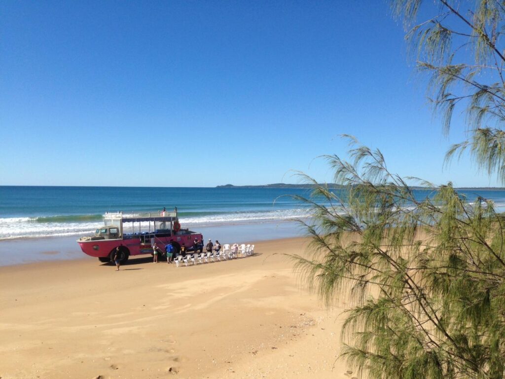 A red amphibious vehicle is parked on a sandy beach near the water, with several people and white chairs arranged in front of it under a clear blue sky.