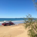 A red amphibious vehicle is parked on a sandy beach near the water, with several people and white chairs arranged in front of it under a clear blue sky.