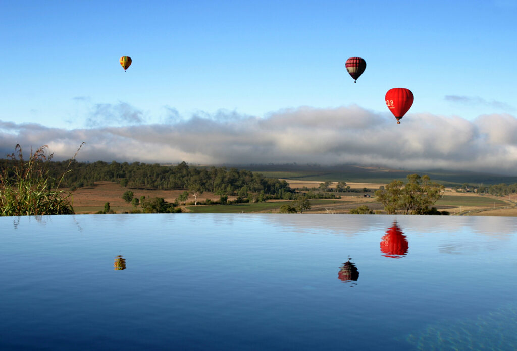 Three hot air balloons float above a rural landscape with trees and low clouds, reflected in a calm body of water in the foreground.