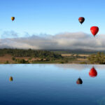 Three hot air balloons float above a rural landscape with trees and low clouds, reflected in a calm body of water in the foreground.