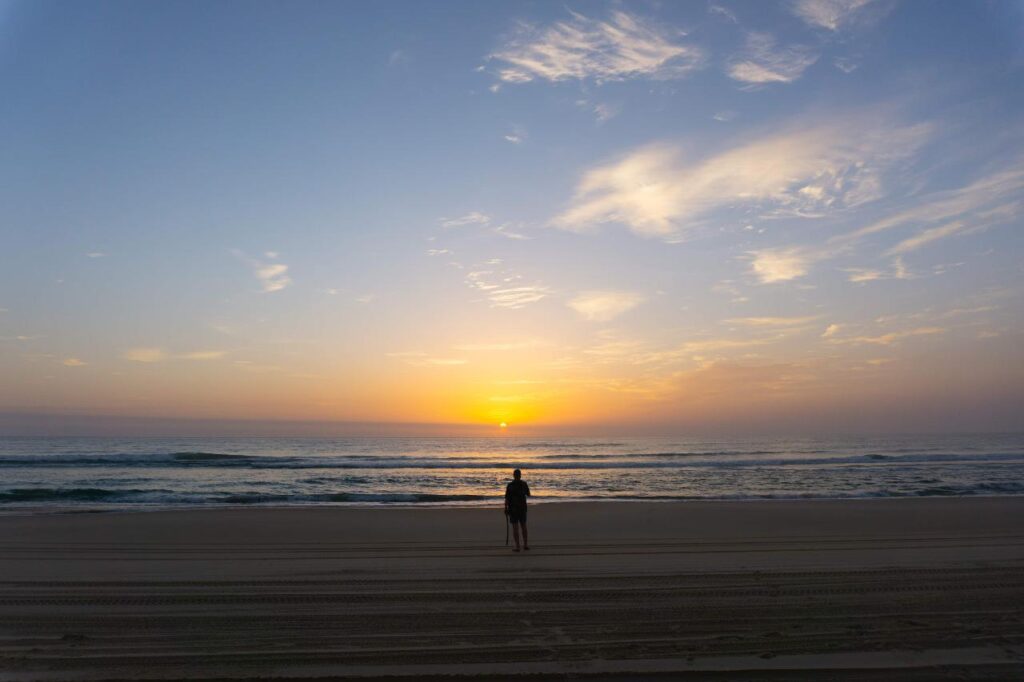 A person stands alone on a sandy beach facing the ocean, watching the sun rise over the horizon under a partly cloudy sky.