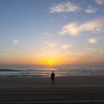 A person stands alone on a sandy beach facing the ocean, watching the sun rise over the horizon under a partly cloudy sky.