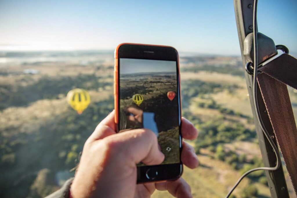 A person in a hot air balloon holds a smartphone, capturing a photo of two other hot air balloons floating over a scenic landscape.